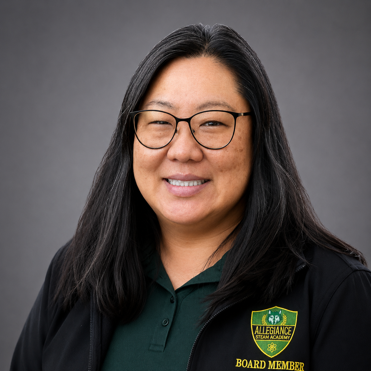 A woman with long dark hair is smiling at the camera. She is wearing a green polo shirt and a black jacket with a logo that says Allegiance STEAM Academy Board Member.